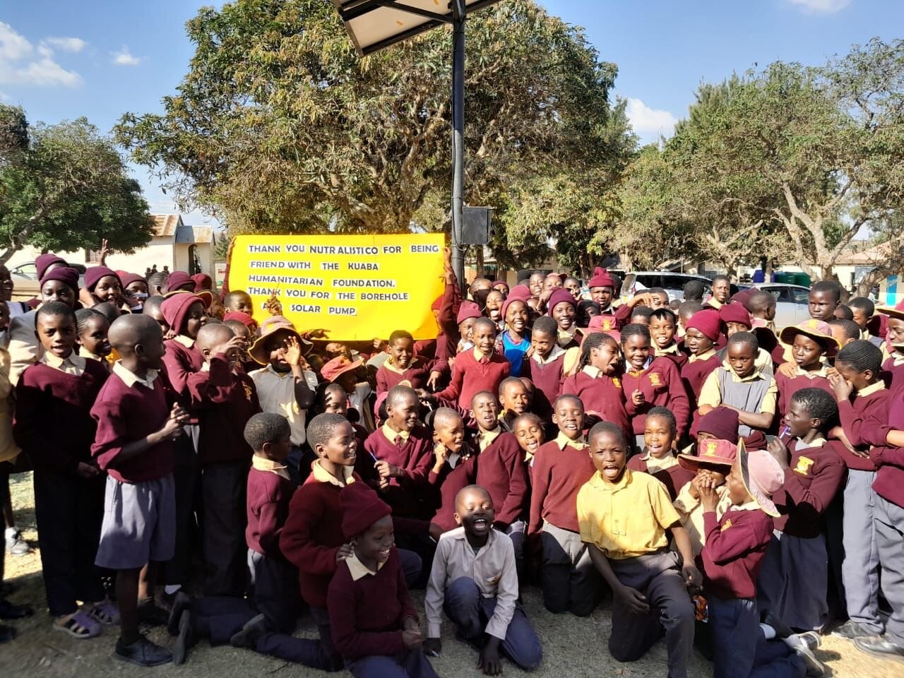 Students from a KHF-supported school in Zimbabwe holding a thank-you banner under a tree, after the borehole and solar pump install